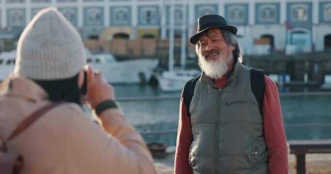 Man, elderly and photo as tourist pose on dock by sea, building and boat on vacation, holiday or travel. Smile, asian and travelling in retirement with photographer for portrait by water in Cape Town