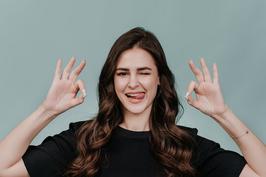 Playful Hispanic Girl With Brown Eyes And Wavy Hair, Blinking, Shows Her Tongue And Gesturing Ok Sign By Both Hands Over Turquoise Studio Backdrop. Everything Is Ok, Success And Happy People Concept.