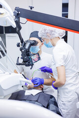 A dentist treats teeth using a dental microscope and tools. The assistant holds a syringe with air...