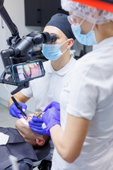 A young dentist looks at the patient's teeth with a dental microscope and holds dental instruments near the mouth. An assistant helps the doctor. They wear white uniforms with masks