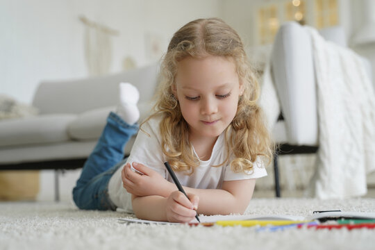 Little caucasian preschooler child girl drawing with crayon, lying on warm floor. Creative art hobby