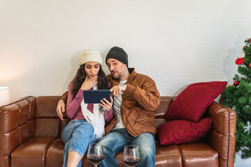 European couple man and woman smiling together looking pointing at tablet mobile device sitting on a brown leather sofa in front of white wall. Male and female wearing warm winter clothing.