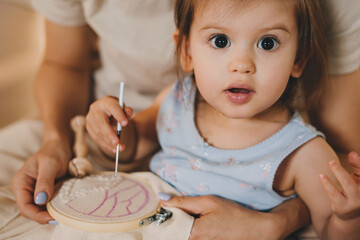 Mother learning baby girl how to embroider a pattern on the white material. Little girl with...