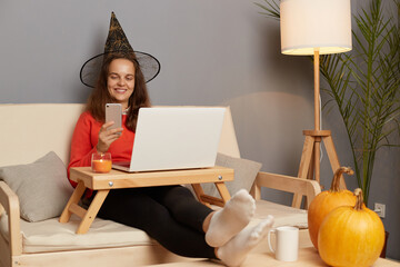 Indoor shot of attractive caucasian woman wearing witch hat working on laptop in home interior and...