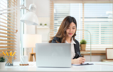 Attractive asian businesswoman using laptop and making notes on notebook.