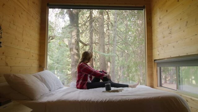 Young woman relaxing in a mountain chalet in winter time. Cinematic dolly shot of beautiful female traveler sitting on bed inside cozy comfortable cabin, observing beautiful dense spruce pine forest
