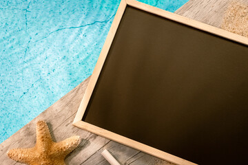 Tableau en ardoise sans inscription vu du dessus sur un dallage en bois au dessus d'une piscine avec une étoile de mer. Ambiance vacances en été.