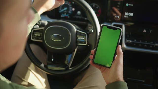 A man holds in his hand a mobile phone with a green screen sitting in a car
