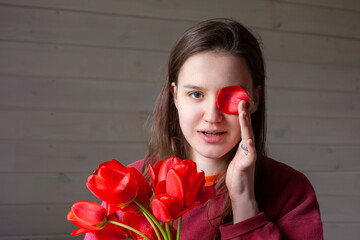 portrait of a brown-haired woman with a bouquet of red tulips on a gray wooden background. a girl in a red pullover with long hair holds a bouquet of tulips in her hands and fools around. Copy space. 