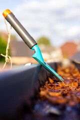 A close up portrait of a roof gutter full of fallen autumn leaves on a sunny day with a small blue garden shovel in it of someone who is cleaning the gutter during the fall season annual chore.