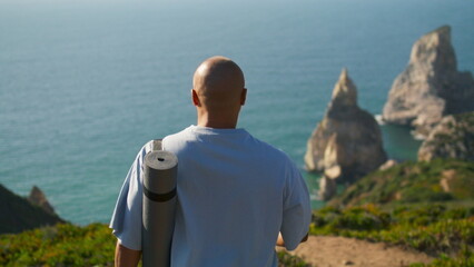 Fototapeta premium Yoga man walking edge cliff holding mat. Focused sportsman looking ocean coast