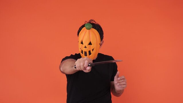 Person with pumpkin mask celebrating Halloween, holding a knife, on orange background. Concept of celebration, All Souls' Day and All Saints' Day.	
