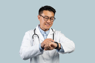 Cheerful middle aged chinese man doctor in white coat, glasses looks at watch on hand, isolated on blue background