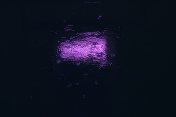 It is raining in a downtown city location. Reflection of purple neon coloured lights from a puddle on the city pavement