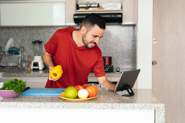 Latin man consulting an online recipe with a tablet while preparing a meal in his home kitchen.
