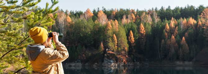 Young woman in yellow hat looking through binoculars at birds on lake against autumn forest...