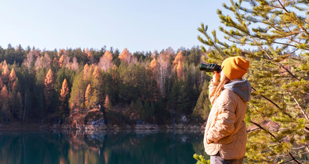 Young woman in yellow hat looking through binoculars at birds on lake against autumn forest Birdwatching, zoology, ecology. Research in nature, observation of animals Ornithology copy space