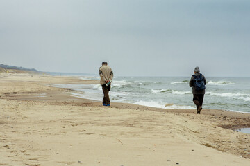 Couple of elderly people walking on the shore of a sandy beach with beautiful rough grey sea with waves, dramatic cloudy grey sky in winter