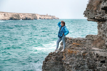 A woman in a blue jacket stands on a rock above a cliff above the sea and looks at the raging ocean. Girl traveler rests, thinks, dreams, enjoys nature. Peace and calm landscape, windy weather.