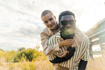 Stunning outdoor portrait of multi-ethnic couple enjoying time outdoors. Handsome tall black man in sunglasses smiling at camera, taking his caucasian bald girlfriend on piggyback. High quality photo