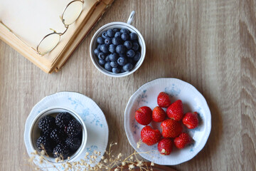 Vintage tea set filled with blueberries, blackberries and starwberries, open book, reading glasses and decorative flowers on the table. Flat lay.