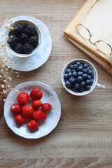 Vintage tea set filled with blueberries, blackberries and starwberries, open book, reading glasses and decorative flowers on the table. Flat lay.