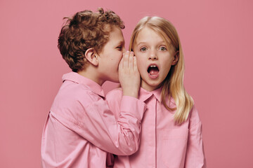 cute, beautiful children are standing in pink clothes on a pink background and the boy tells the girl secrets in her ear. Topics of friendship and relationships