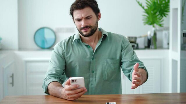 Dark-haired bearded Hispanic young man in casual shirt, using smartphone, transfers money. He is anxious, frustrated not paying the bill due to lack of coins, throws the credit card on a kitchen table