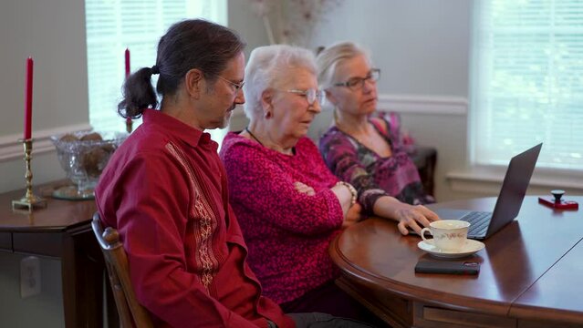 Elderly senior mother with mature son and daughter in law in dining room sitting in front of laptop computer looking at photos and talking about memories.