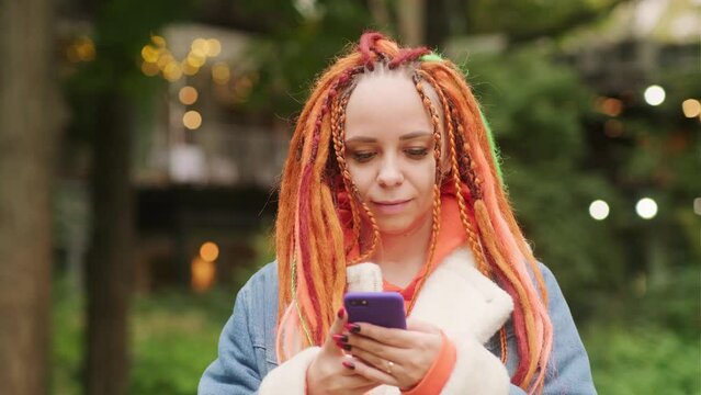 Young woman with dreadlocks browsing mobile phone, standing in romantic parkland. Pretty female with colourful multicoloured hairstyle using smartphone.