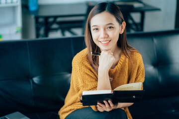 Portrait of a smiling Asian woman sitting on a sofa. Using laptop in the living room at home, working at home via online video conference.