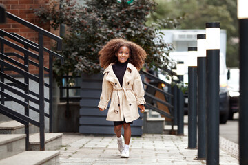 Little African American girl with long curly hair walking city street. Happy child smiling, wearing...