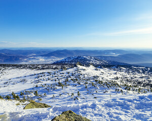 View from mount peak to mountain hills, white snow, blue sky, stones and fir trees. Winter landscape in Gornaya Shoria, Sheregesh ski resort in Russia, Altai mountains nature background.