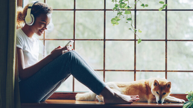 Pretty young African American girl is listening to music with headphones and using smartphone sitting on window sill with lovely well-bred dog lying near her.
