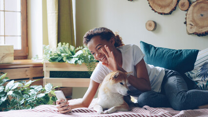 Emotional African American girl is talking on mobile phone making video call smiling and chatting looking at screen with obedient pedigree dog lying on bed at home beside her. - Powered by Adobe