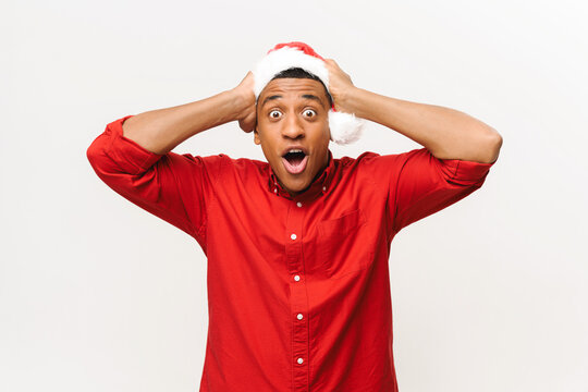 Shocked African-american Guy In Santa Hat And Red Shirt Holding Head With Both Hands Isolated On White Background, Astonished Guy Looks At Camera With Wide Opened Eyes And Mouth, Incredible Promotion