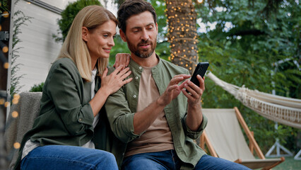 Married couple husband and wife Caucasian bearded man and blond woman 40s spouses outdoor in backyard at campsite looking at phone choose buy online booking with smartphone using mobile e-shopping app
