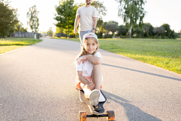 child girl sitting down on a skateboard and having his father push her.