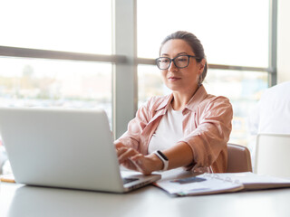 Woman in eyeglasses works with laptop. Modern office with panoramic windows at co-working center. Workplace for freelancers in business center.