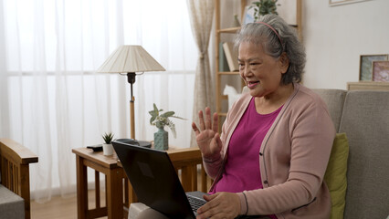 Fototapeta na wymiar happy Asian senior woman sitting on the sofa and waving hi to the screen while making a video call on the laptop to her child in the living room at home