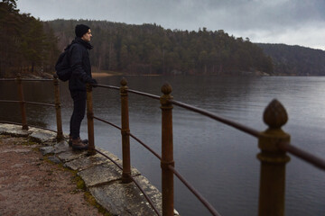 A caucasian man with a backpack standing by a old fence looking out over a lake at a rainy day.