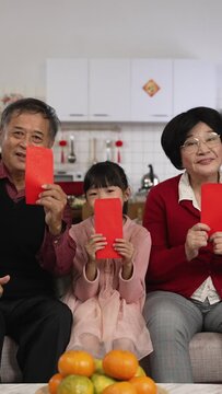 Vertical Screen: generation smiling at camera with thumb and victory hand gestures while taking burst photos with red envelopes lucky money at home on chinese lunar new year.  translation: fortune