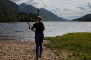 A woman plays the Scottish bagpipes on the beach at the Loch Shiel lookout, next to the Glenfinnan Monument - National Trust for Scotland