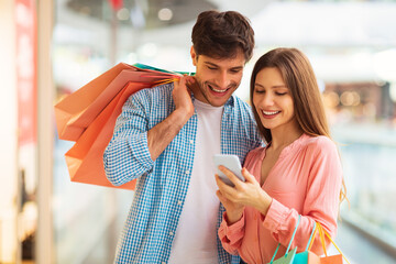 Couple Shopping Online Using Smartphone Posing Holding Bags In Mall