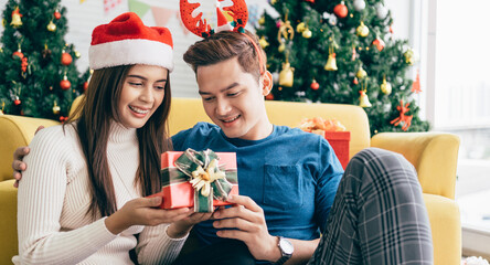 Young beautiful happy Asian woman wearing Santa Claus hat surprises her boyfriend with a Christmas gift at home with Christmas tree in the background. Image with copy space.