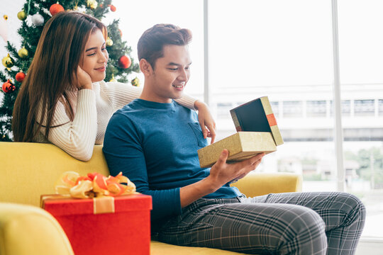 Young beautiful happy Asian woman surprises her boyfriend with a Christmas gift at home with Christmas tree in the background. Image with copy space.
