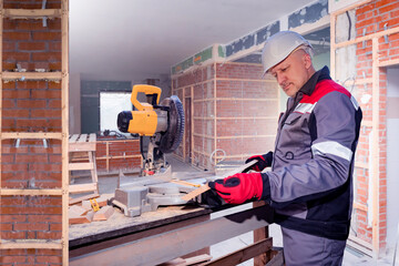 Construction works. Builder man next to machine for sawing. Builder stands at workplace. Guy in construction uniform. Builder uses circular stationary saw. Man in building under construction