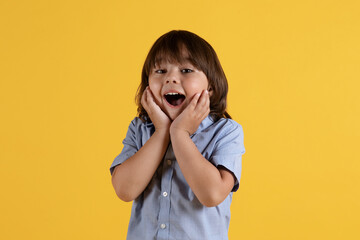 No way. Surprised little boy touching his cheeks in amazement, looking at camera with open mouth, orange background