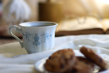 Cup of tea or coffee, plate of chocolate chip cookies, open book, reading glasses, candle and vase with flowers. Selective focus.