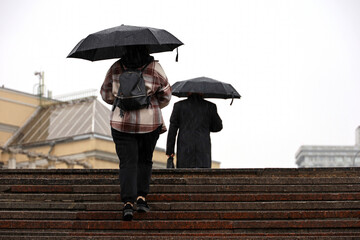 People walking up the steps on city buildings background, woman with umbrella in foreground. Heavy rain in autumn city